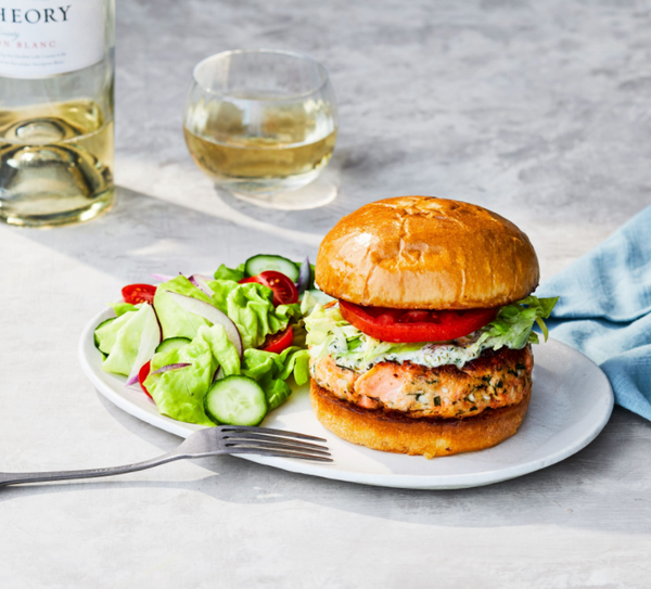 Salmon burger with lettuce, tomato, sauce, side salad, fork, and glass of white wine on a light surface
