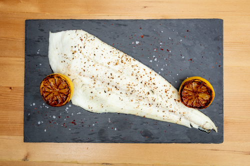 Cooked halibut fillet seasoned with pepper and salt flakes, served with two grilled lemon halves on a slate board.