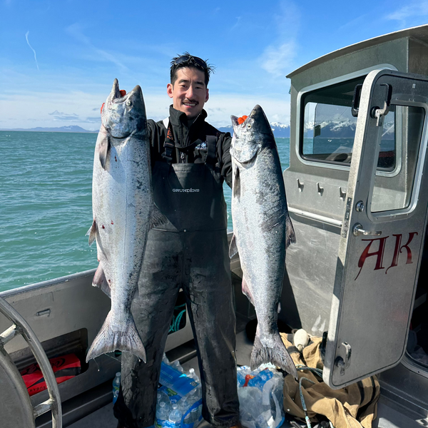 Person on a fishing boat holding two Copper River king salmon; boat door shows letters AK