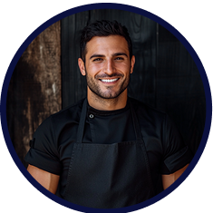 Smiling man wearing a black apron and black shirt, standing in front of a dark background.