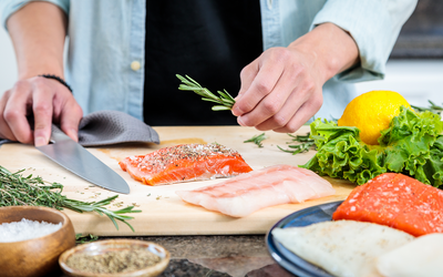 Hands seasoning raw fish fillets on a wooden cutting board with herbs, lemon, knife, and spice bowls.