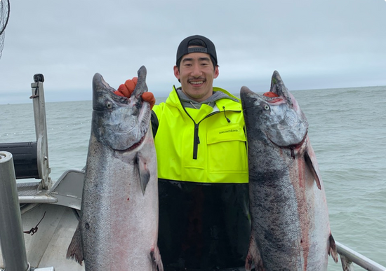 Smiling man in neon yellow rain jacket and black hat holding two large salmon side-by-side on a fishing boat at sea.