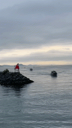 Person in a red jacket sitting on rocks by the water with boats in the distance under a cloudy sky.