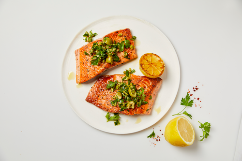 Two seared salmon fillets on a white plate topped with herb sauce, with grilled lemon halves and parsley garnish.
