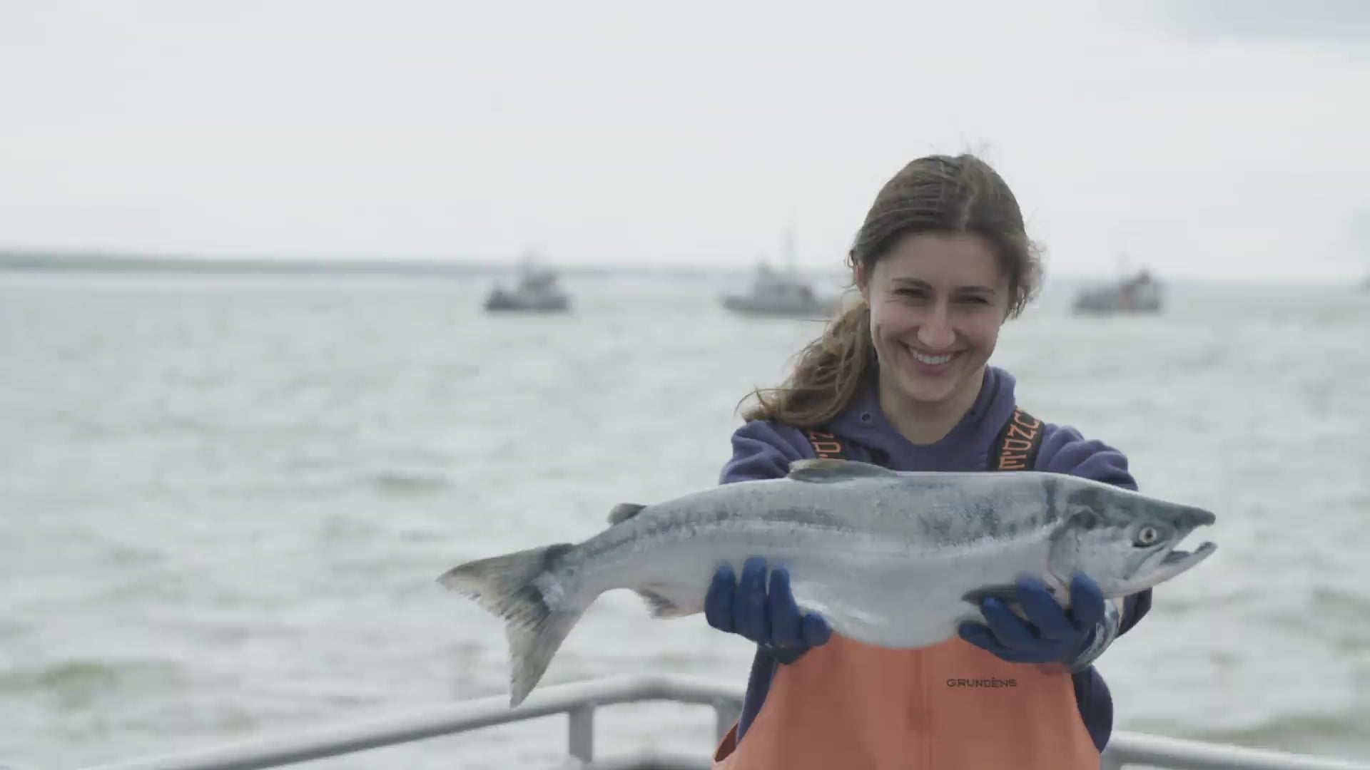 Smiling woman on a boat holding a large salmon, wearing an orange bib labeled 'GRUNDENS'.