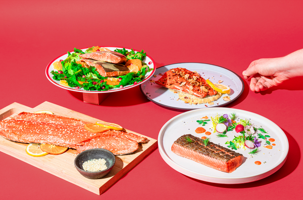 Four plated Copper River salmon dishes on a red surface, with a hand reaching toward one plate
