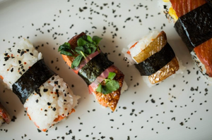 Three salmon musubi pieces wrapped in nori on a white plate sprinkled with black sesame