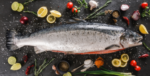 Whole raw salmon with lemon slices, cherry tomatoes, garlic, herbs, and spices on dark surface.