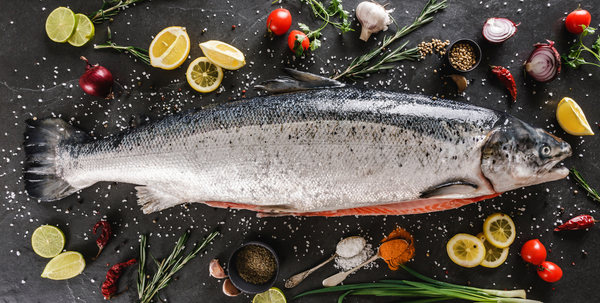 Whole raw salmon with lemon slices, cherry tomatoes, garlic, herbs, and spices on dark surface.