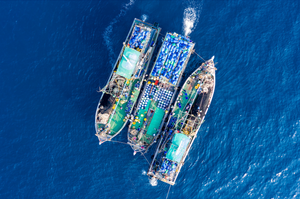 Aerial view of three fishing boats tied side-by-side, decks full of blue barrels and fishing gear on deep blue ocean.