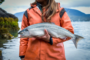 Person in orange rain jacket holding a large silver salmon by the water with mountains in the background