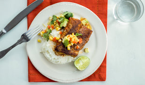 Blackened Wild Alaskan cod on rice with avocado corn salsa and lime on white plate over red napkin, silverware and water glass