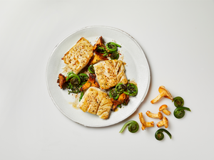 Three seared white fish fillets on a white plate with chanterelle mushrooms, fiddlehead ferns, and a light sauce.