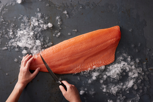 Hands using a knife to remove pin bones from a salmon fillet on a dark surface with crushed ice