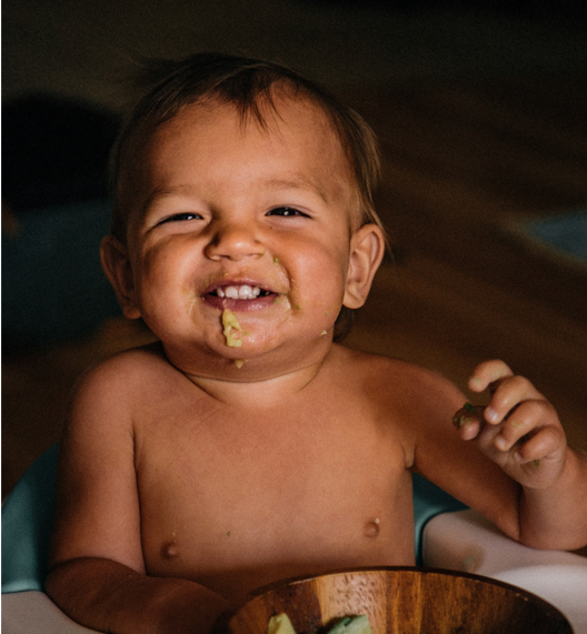 Smiling baby with food on face sitting in a high chair holding a piece of food near a wooden bowl.