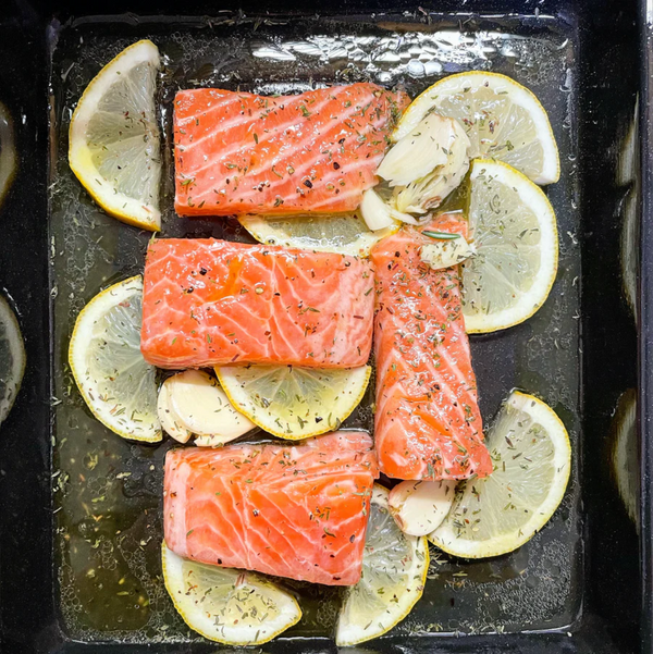 Raw salmon fillets seasoned with herbs, lemon slices, and garlic in a black baking tray.