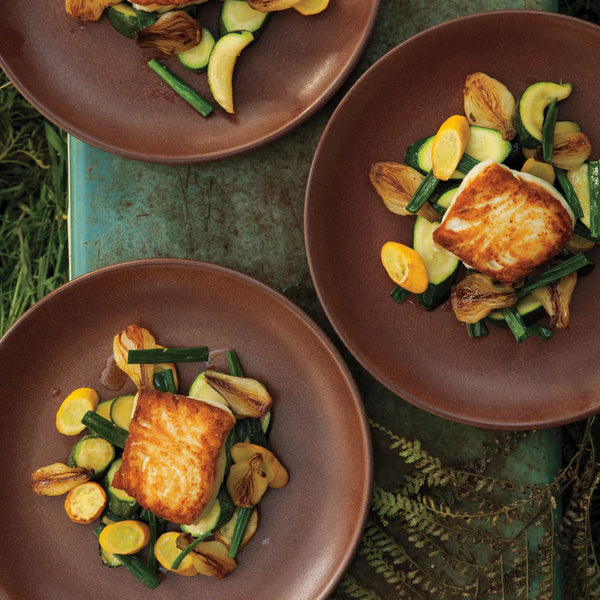 Three brown plates each with a cooked halibut fillet and sautéed vegetables including zucchini, yellow squash, and onions