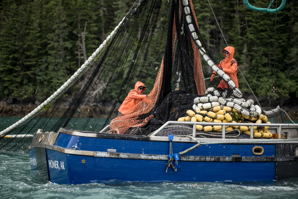Two people in orange rain gear handling fishing nets on a blue fishing boat labeled 'HOMER, AK'