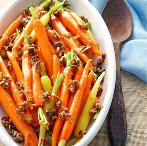 Glazed carrots with chopped nuts in a white dish next to a wooden spoon on a blue cloth