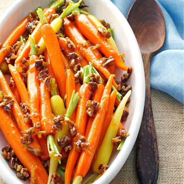 Glazed carrots with chopped nuts in a white dish next to a wooden spoon on a blue cloth