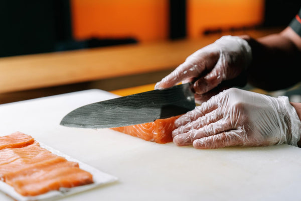 Hands in gloves slicing salmon with a large knife on a white cutting board