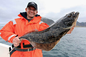 Smiling man in an orange waterproof jacket holding a large halibut on a boat, with mountains and cloudy sky in background