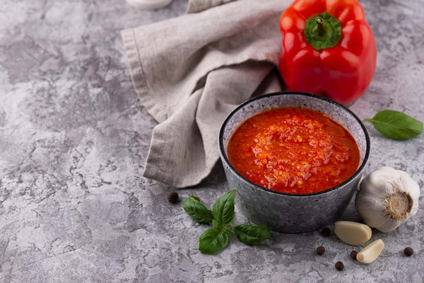 bowl of red marinade sauce with garlic cloves, basil leaves, and red bell pepper on gray surface