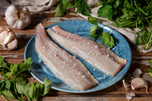 Two salted cod fillets on a blue plate sprinkled with coarse salt, garlic cloves and parsley sprigs nearby.
