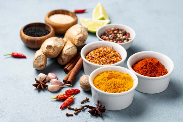 Various spices and ingredients in bowls including turmeric, chili powder, crushed red pepper, cinnamon sticks, garlic, ginger, star anise, cloves, and lime wedges.