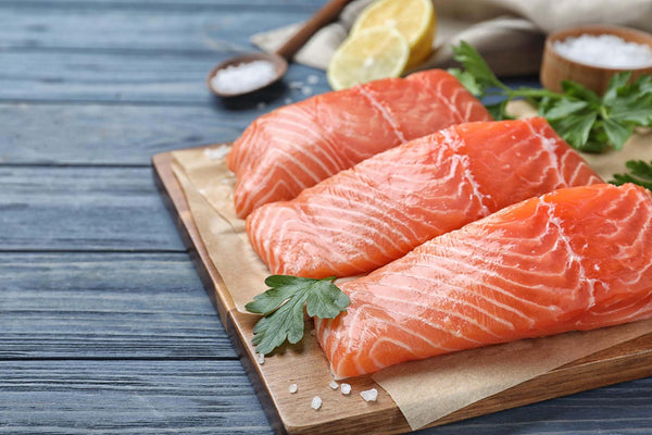 three raw salmon fillets on a wooden board with lemon slices, parsley, and coarse salt