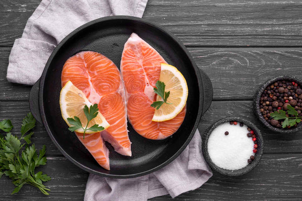 Two raw salmon steaks with lemon slices and parsley in a black pan, next to salt and pepper bowls on a wooden surface.