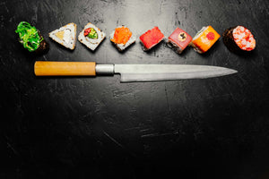 long sashimi knife with wooden handle below assorted sushi rolls arranged across a black slate surface