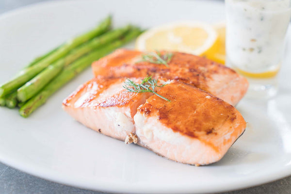 Two cooked salmon fillets garnished with dill, served with asparagus and lemon slices on a white plate.