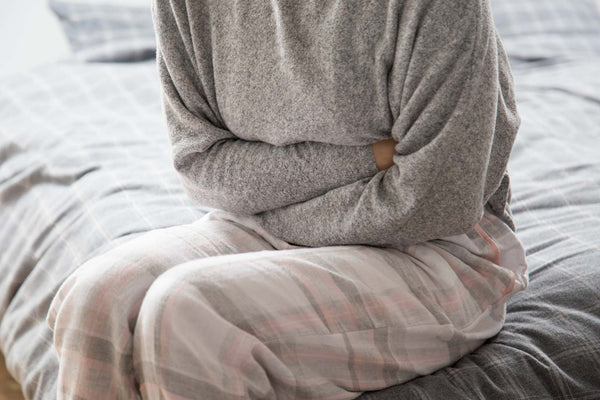 Person clutching abdomen, sitting on a bed, possibly indicating stomach pain or discomfort