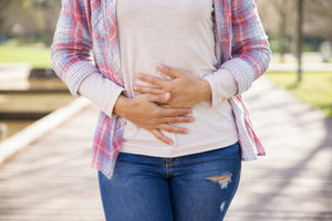 Woman standing on a boardwalk clutching her lower abdomen, wearing a plaid shirt and ripped blue jeans.