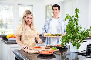Pregnant woman seasoning salmon on a kitchen island while partner smiles and holds a tray of salad greens.