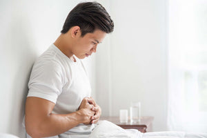 Young man sitting on a bed clutching his abdomen in pain; glass of water and pill bottle on a nearby nightstand.