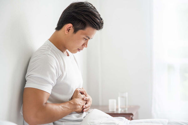 Young man sitting on a bed clutching his abdomen in pain; glass of water and pill bottle on a nearby nightstand.