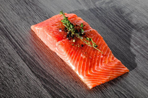 Raw king salmon fillet on dark wooden surface garnished with thyme sprig and cracked black pepper.
