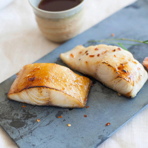 Two seared cod fillets with glaze and red pepper flakes on a slate serving board, ceramic cup blurred in background