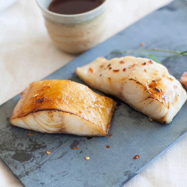 Two seared cod fillets with glaze and red pepper flakes on a slate serving board, ceramic cup blurred in background