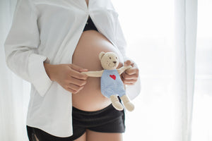 Bare pregnant belly with hands holding a small teddy wearing blue-striped outfit with red heart