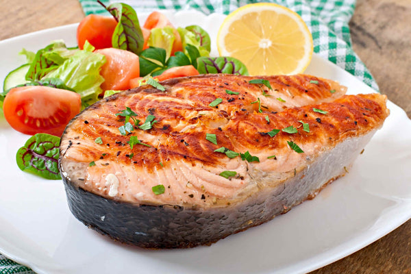 Grilled salmon steak garnished with parsley, served with salad and a lemon half on a white plate.