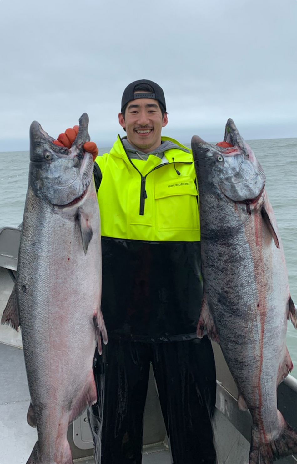 Man in yellow rain jacket holding two large fish on a boat at sea under cloudy sky